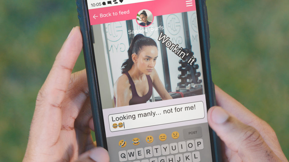 A photo of a young woman is shown on a phone where someone is making a negative comment underneath. She is sitting on a bench in a gym in front of a wall lined with mirrors. Her facial expression shows her concentrating hard as she lifts weights. The comment under the photo says 'Looking manly, not for me' followed by two laughing face emoji's.