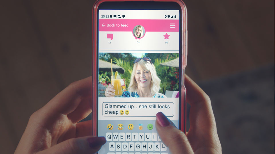 A photo of a middle aged white woman, smiling and holding up a drink outside at a beach bar is shown on a phone where someone is making a negative comment underneath. The comment under the photo says 'Glammed up, she still looks cheap' with two crying with laughter emojis.