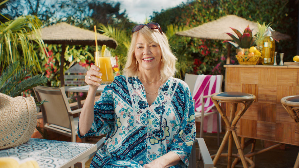 A middle aged white woman is smiling and holding up a drink as she sits outside at a beach bar, surrounded by tropical plants. There is a bar to the right of the women, and other tables, chairs and sun umbrellas behind her.